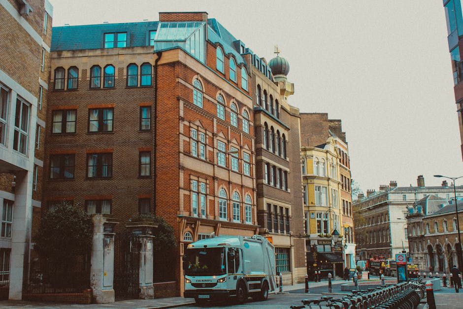 A small light blue waste collection vehicle parked on a city street appears to be involved in a rubbish removal or on-site clearance operation. The vehicle is positioned in front of a row of historic 