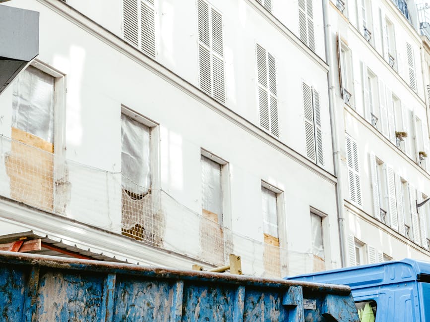 A small light blue waste collection vehicle parked on a city street appears to be involved in a rubbish removal or on-site clearance operation. The vehicle is positioned in front of a row of historic 