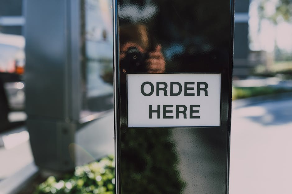 A close-up view of a black metal stand with a white rectangular sign that reads 'ORDER HERE' in bold black letters, set outdoors in a parking area. In the background, blurred images of a silver or gre
