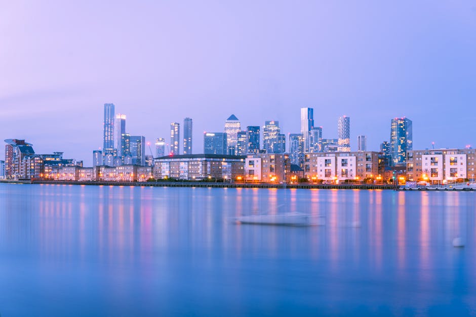 A panoramic view of a modern city skyline showcasing a variety of tall commercial and office buildings with glass and steel facades, some featuring geometric patterns and reflective surfaces. The fore